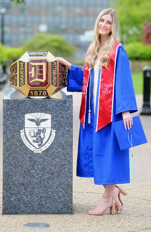 Abby Leister in graduation regalia with hand on Duquesne ring monument and green campus trees and city buildings in background.